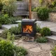Barbecue à bois Hestia 300 Heat & Grill avec foyer vitré allumé, installé près d’un muret dans un jardin